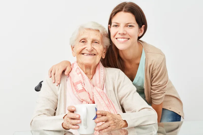 Nahaufnahme einer lächelnden Seniorin, die von einer jungen, lächelnden Pflegekraft umarmt wird.