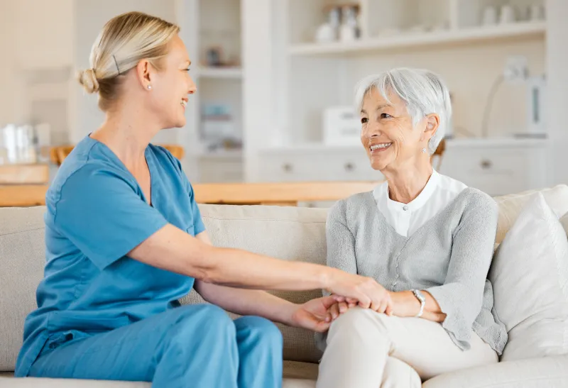 Eine lächelnde Pflegekraft im blauen Kasack hält im Gespräch auf einem Sofa die Hand einer lächelnden Seniorin.