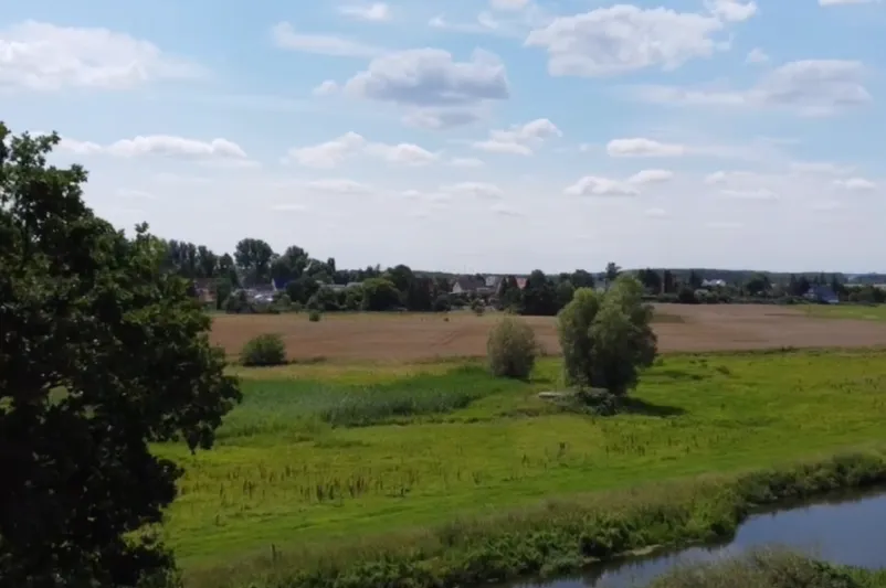 Weitläufige ländliche Landschaft mit grünen Feldern, einigen Bäumen, einem Fluss im Vordergrund und vereinzelten Häusern am Horizont unter einem bewölkten Himmel.