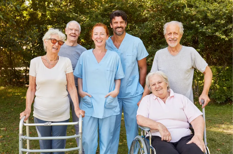 Eine lächelnde Gruppe aus vier Senioren und zwei Pflegekräften steht oder sitzt draußen in einer Grünanlage.